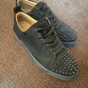 Louboutin svart mocka - Säljer ett par svarta sneakers från Christian Louboutin med coola svarta nitar på tån och klassisk röd sula. Skorna har snörning, rund tå och är gjorda i mocka med skinnfoder. Finns låda osv, ligger extrasulor i just nu som ni får med