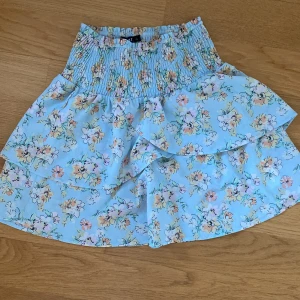 Blommig volangkjol från LMTD - Supersöt ljusblå kortkjol från LMTD med blommigt mönster i pastellfärger. Kjolen har bred smock i midjan och dubbla volanger som ger en flowy känsla. Perfekt för vår och sommar när du vill ha en fräsch och lekfull look.