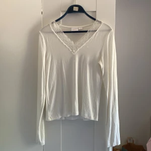 Vit långärmad topp med spets från Gina Tricot - Säljer en vit långärmad topp från Gina Tricot i mjuk bomull. Toppen har en snygg v-ringning med spetsdetaljer som ger en romantisk vibe. Passar perfekt till jeans eller kjol och är superenkel att styla. Spetsen är vit och har ett blommigt mönster.