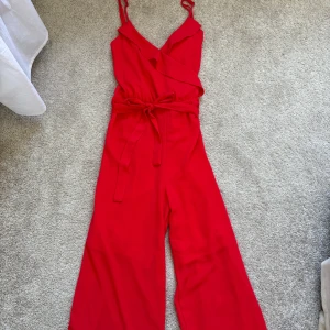 Röd byxdress med omlott och knytband - Snygg röd byxdress med omlott upptill, smala axelband och ett matchande knytband i midjan. Vida ben ger en cool och avslappnad vibe. Perfekt för dig som vill sticka ut med färg och stil. Materialet känns mjukt och följsamt mot kroppen.  Storlek 36❤️