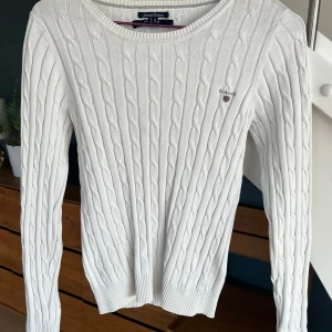 Vit kabelstickad tröja från GANT xs - Säljer en vit/krämvit kabelstickad tröja från GANT i stretchig bomull. Tröjan har rund hals, ribbade muddar och lång ärm. GANT-loggan är broderad på bröstet. Perfekt för dig som gillar klassisk och clean stil.