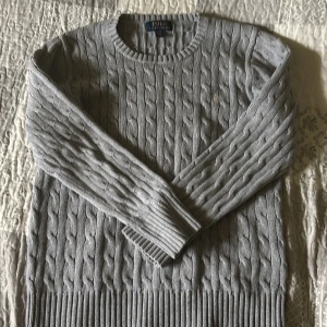 Grå kabelstickad tröja Polo Ralph Lauren - Säljer en snygg grå kabelstickad tröja från Polo Ralph Lauren. Tröjan har rund hals, långa ärmar och klassisk ribbad kant nedtill och vid ärmslut. Perfekt för dig som gillar stilrena och tidlösa plagg med en preppy vibe.