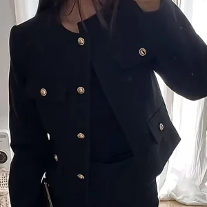 Svart kavaj med guldknappar - Snygg svart kavaj med guldiga knappar framtill och på fickorna. Modellen är croppad och har en stilren, rak passform med långa ärmar. Perfekt för dig som gillar klassisk men trendig stil. Materialet känns som polyester och kavajen har en elegant look.