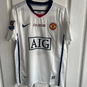 Manchester United Rooney 10 Final Roma 2009 tröja - Säljer en vit Manchester United fotbollströja från Nike med tryck 'Final Roma 2009' och AIG sponsor på bröstet. Tröjan har Wayne Rooney och nummer 10 på ryggen, samt mörkblå detaljer längs sidorna och kragen. Materialet är lätt och andas, perfekt för match eller träning.
