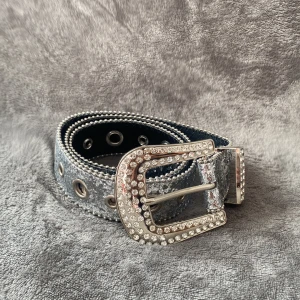 Glittrigt silverbälte med strass - Säljer ett snyggt bälte i silver med glittrig yta och stora metallhål längs hela bandet. Spännet är dekorerat med massor av strass som ger extra bling. Perfekt för dig som vill sticka ut och addera lite edge till din outfit.