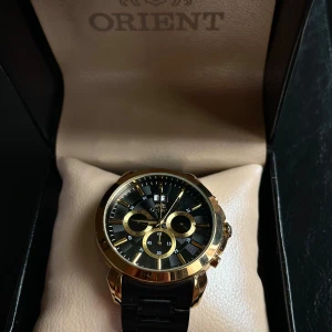 Svart och guld kronografklocka Orient - Pris kan diskuteras.                                             ordinarie pris:6,300kr.                                          Snygg kronografklocka från Orient med svart urtavla och guldiga detaljer. Boetten är i rostfritt stål med guldfinish och armbandet är svart. Klockan har tre subdials, datumfönster och stilrena indexmarkeringar. Kommer i original ask.