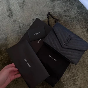 Svart axelväska från Saint Laurent - Säljer en svart axelväska från Saint Laurent med klassiskt YSL-emblem framtill. Väskan har en elegant, strukturerad yta med chevronmönster och kedjerem. Perfekt för dig som vill ha en stilren och lyxig accessoar.
