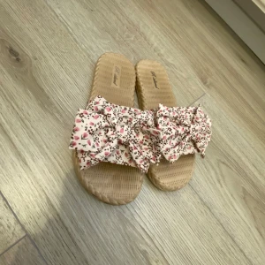 Blommiga sandaler med rosett - Säljer ett par söta sandaler med beige sula och stor rosett framtill i blommigt tyg i rosa och vitt. Perfekta för varma dagar och ger en somrig känsla till outfiten.Dom är från Kroatien 🇭🇷 