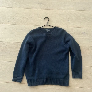 Mörkblå stickad tröja från Polo Ralph Lauren - Mörkblå stickad tröja från Polo Ralph Lauren med diskret broderad logga på bröstet. Klassisk modell med rund halsringning och ribbade muddar vid ärmslut och nederkant. Perfekt för lager-på-lager under kyliga dagar.