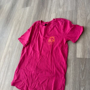 Röd t-shirt med SUN DAY-tryck - Cool rosa t-shirt med trycket 'SUN DAY' och en sol på bröstet. Klassisk passform och rund halsringning. Perfekt för dig som vill ha en färgstark och enkel look.