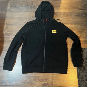 Svart hoodie med dragkedja från Hugo - Säljer en svart hoodie från Hugo med dragkedja framtill och huva med snörning. På bröstet finns en gul patch med Hugo-loggan. Perfekt för en avslappnad och stilren look.
