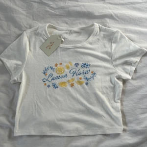Aurelle Baby Tee - Oanvänd Baby tee från Aurelle med trycket ”Lemon Flora”. Super skönt material! Står att det är storlek M men passar mer som en XS-S!