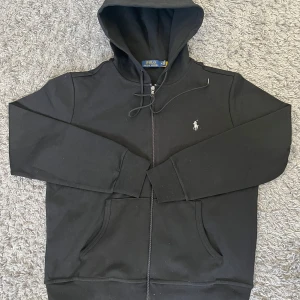 Svart hoodie med zip från Ralph Lauren - STORLEK M. Snygg svart hoodie från Ralph Lauren med dragkedja och broderad logga på bröstet. Hoodien har luva och två fickor framtill. Perfekt för en avslappnad och stilren look. Nästan helt oanvänd och helt ren. PRISET KAN DISKUTERAS