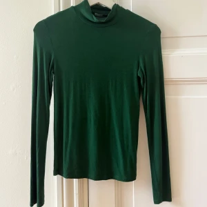 Grön polotröja från Stradivarius - Mörkgrön polotröja från Stradivarius i mjukt material. Tröjan har lång ärm och en smal passform som sitter snyggt mot kroppen. Perfekt att styla med jeans eller kjol för en enkel och stilren look.