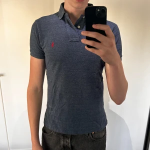 Blå pikétröja från Ralph Lauren - Säljer en klassisk blå pikétröja från Ralph Lauren med röd broderad logga på bröstet. Tröjan har korta ärmar, krage och knappar vid halsen. Perfekt för en stilren och avslappnad look. Använd men bra skick! Storlek S