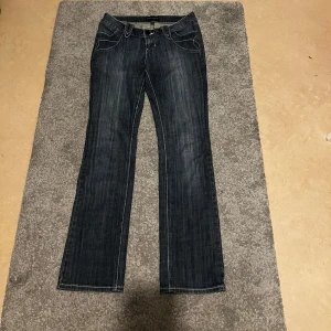 Mörkblå bootcut jeans från Jean Genie - Snygga mörkblå jeans från Jean Genie med bootcut passform. Jeansen har dekorativa detaljer på bakfickorna, bland annat en dragkedja. Klassisk femficksmodell med vita kontrastsömmar och låg midja.