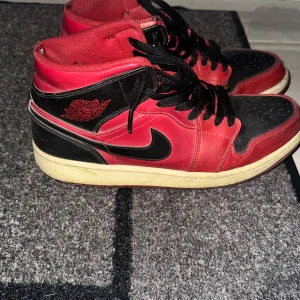 Nike Air Jordan 1 Mid  - Säljer ett par klassiska Nike Air Jordan 1 Retro High i rött och svart med svarta skosnören och vit sula. Skorna har den ikoniska Jordan-loggan på sidan och är tillverkade i skinn. Perfekta för dig som gillar streetwear och sneakerskultur. Behöver bara en tvätt sen är de redo att rocka.