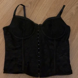 Svart korsett-topp med snörning - Säljer en svart korsett-topp med justerbara axelband och hakknäppning framtill. Toppen har snörning i ryggen som gör att man kan justera storleken mycket. Den är storlek S men skulle säga att den passar både s och xs då man kan dra åt den väldigt mycket om man vill. Den är endast testad. Den har inga hål eller fläckar eller tecken på användning eller något sånt. Den har ej lappen kvar
