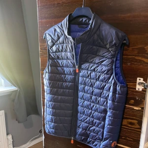 Save the duck väst  - En fin marinblå väst med ljusblå insida och orangea detaljer, mycket bra skick. Medföljer en tillhörande påse. Storlek small, pris 500kr