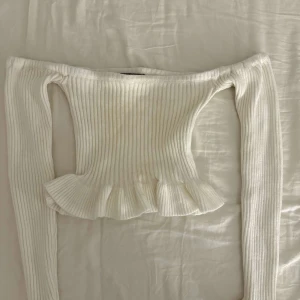 Vit ribbad offshoulder topp med volang - Trendig vit offshoulder topp med ribbad struktur och volang nertill. Långärmad och croppad modell som passar perfekt till jeans eller kjol.