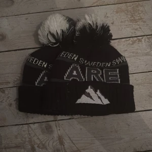 Svart och vit mössa med tofs från Åre - Säljer en svart stickad mössa med vit tofs och texten 'Åre Sweden' runt om. Mössan har ett broderat bergmotiv framtill och är perfekt för kalla dagar.