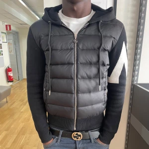 Svart pufferjacka med huva från Moncler - Säljer en svart pufferjacka från Moncler med huva och dragkedja. Jackan har quiltad framsida, stickade ärmar och rygg samt en vit Moncler-logga och mönster på ärmen. Perfekt för kyliga dagar. Hör av er vid minsta lilla intresse. Priset är inte hugget i sten. Använd helst INTE instabox.