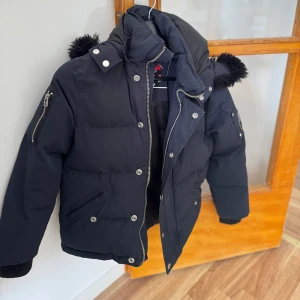 Svart dunjacka från Moose Knuckles - (Pris kan diskuteras) Säljer en svart dunjacka från Moose Knuckles med pälskantad huva och silvriga dragkedjor. Jackan har flera fickor med tryckknappar och dragkedjor samt ribbade muddar vid ärmslut. Perfekt för kyliga dagar. Jack är då väll använd och därför slitningar på märket, jag säljer denna nu efter som den är alldeles för lite för mig. Storlek är XS.  