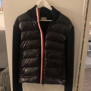 Svart Cardigan från Moncler - Säljer en svart cardigan från Moncler med stickade ärmar och klassisk röd-vit-blå rand vid dragkedjan. Jackan har hög krage och Moncler-logga på ärmen. Är inköpt av en som till 100% garanterar att allt han säljer är äkta. Vid frågor och funderingar, tveka inte på att höra av dig.