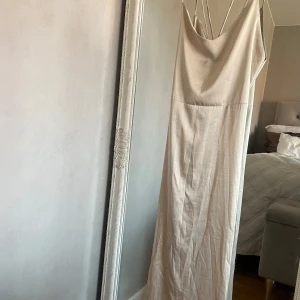 Champagnefärgad midiklänning med snörning - Säljer en elegant champagnefärgad midiklänning med tunna axelband, öppen rygg och snörningsdetalj baktill. Klänningen har dragkedja i sidan och är tillverkad i ett mjukt, glansigt material som ger en lyxig känsla.