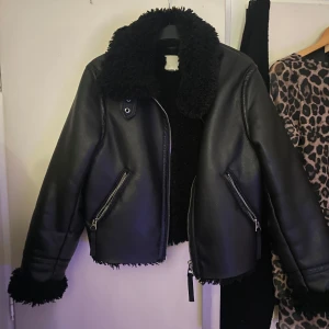 Svart skinnjacka med fuskpälsfoder från H&M - Säljer en svart skinnjacka från H&M med mjukt fuskpälsfoder och krage. Jackan har dragkedja framtill och på fickorna samt detaljer i silver. Perfekt för kyligare dagar och ger en cool look.