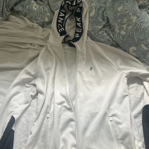 Vit hoodie från Peak Performance med svart huva - Storlek L men passar även M. Säljer en vit hoodie från Peak Performance med svart text på huvan och svarta muddar. Jackan har dragkedja framtill och två fickor med dragkedja. Perfekt för en sportig och avslappnad stil. Orginal pris 1600kr mitt pris 750. Pris går att diskutera! 