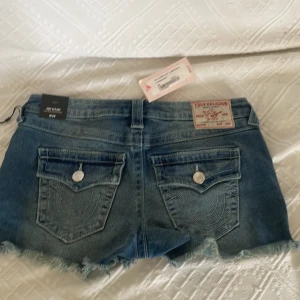 Blå jeansshorts från True Religion - Säljer ett par blå jeansshorts från True Religion med råa fransade benslut och klassiska bakfickor med lock och knapp. Shortsen har normal passform och är tillverkade i denim. Perfekta för sommaren!
