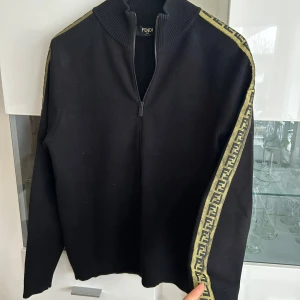 Svart zip-jacka från Fendi med logoband - Svart jacka från Fendi med dragkedja och ribbad krage. Guldig logga längs ärmarna. Nypris: 12 500kr