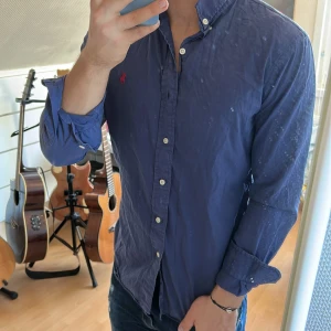 Blå skjorta från Ralph Lauren - Säljer en klassisk blå skjorta från Ralph Lauren med röd broderad logga på bröstet. Skjortan har knappar hela vägen, button-down krage och långa ärmar. Perfekt för dig som gillar stilrena och tidlösa plagg.