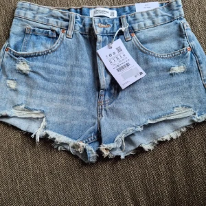 Ljusblå jeansshorts från Pull&Bear - Ljusblå jeansshorts från Pull&Bear med slitna detaljer och fransig kant. Klassisk femficksmodell med hög midja och råa benslut. Perfekta för varma dagar och en avslappnad stil. Helt nya med prislappen kvar!!!!