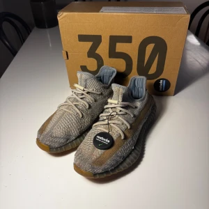 Adidas Yeezy Boost 350 V2 - Säljer ett par Adidas Yeezy Boost 350 V2 med stickad ovandel i beige och grå toner samt genomskinlig sula. Skorna har snörning och den klassiska Yeezy-designen. Perfekta för dig som gillar streetwear och sneakers med unik look.