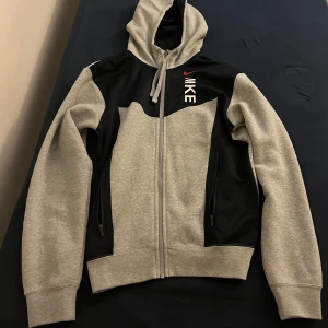 Nike hoodie - Snygg hoodie från Nike i grått och svart med dragkedja och huva. Stora fickor framtill och Nike-logga på bröstet. Perfekt för en sportig och avslappnad stil.