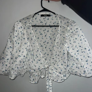 Vit blommig blus med knyt från Bik bok - Supersöt vit blus med små blå blommor, volangdetaljer och knyt framtill. Blusen har trekvartsärmar och är croppad, perfekt för vår och sommar. Tunn och luftig i bomullsmaterial.