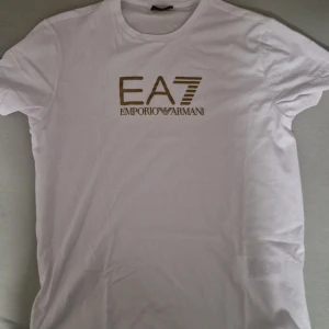 Vit t-shirt från Emporio Armani EA7 - Snygg vit t-shirt från Emporio Armani EA7 med guldig logga tryckt på bröstet. Klassisk rund halsringning och korta ärmar. Tillverkad i mjuk bomull för en skön känsla hela dagen.