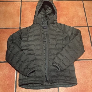 Grön dunjacka med huva från Peak Performance - Säljer en grön dunjacka från Peak Performance med huva och dragkedja framtill. Jackan är quiltad och har långa ärmar samt resår vid ärmslut. Perfekt för kyligare dagar och enkel att matcha med olika outfits. Använd väldigt kort tid, mycket fint skick. 