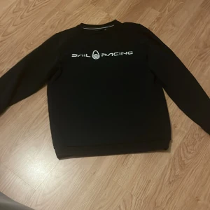 Svart sweatshirt från Sail Racing - Svart sweatshirt från Sail Racing med vit logotyp på bröstet. Tröjan har rund hals och långa ärmar, perfekt för en sportig och stilren look. Passar dig som gillar enkel och klassisk design.