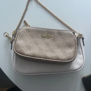 Guldig och vit axelväska från Guess - Snygg axelväska från Guess med två fack, ett i guld och ett i vitt. Väskan har guldfärgad kedja och Guess-logga framtill. Perfekt för att bära det viktigaste med stil.