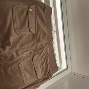 Beige mjukisbyxor från Juicy Couture - Säljer ett par beige mjukisbyxor från Juicy Couture med bred resår i midjan, snörning med gulddetaljer och stora fickor med lock och guldknappar. Mjukt material och klassisk logga broderad på fickan.