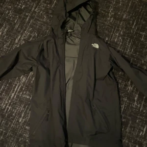 Svart regnjacka från The North Face - Säljer en svart regnjacka från The North Face med huva och dragkedja framtill. Jackan har två fickor med dragkedja och logga på bröstet. Perfekt för blöta dagar och enkel att matcha med allt. Kom gärna med prisförslag 