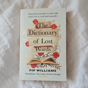 The Dictionary of Lost Words - En roman på engelska med titeln 'The Dictionary of Lost Words' av Pip Williams. Omslaget är ljust med blommiga illustrationer och en öppen bok. Boken handlar om språk, kvinnors röster och historiska förändringar kring Oxford English Dictionary.