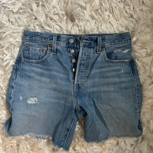 Levi's jeansshorts med slitningar  - Säljer ett par snygga klassiska blå jeansshorts från Levi's med hög midja och knappar framtill. Shortsen har råa kanter och slitna detaljer. Säljer då de är för små för mig. Svårt att säga storleken men jag skulle säga att de passar S och XS.