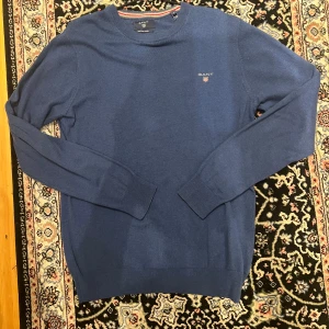 Blå tröja från Gant - Säljer en klassisk blå tröja från Gant med rund halsringning och diskret broderad logga på bröstet. Tröjan är tillverkad i en mjuk bomulls- och ullblandning och har långa ärmar samt ribbade muddar vid ärmslut och nederkant.