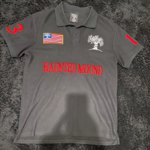 Haunted mound polo svart - Svart pikétröja med röd text 'HAUNTED MOUND' på bröstet, broderad amerikansk flagga och trädmotiv fram och bak. Röda siffror på ärmarna och klassisk krage med knappar. Perfekt för dig som gillar streetwear med unik design.