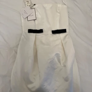 Vit axelbandslös klänning med svarta rosetter från Vila - Säljer en elegant vit axelbandslös klänning från Vila med två svarta rosetter framtill. Klänningen har en slät satinliknande yta och veckade detaljer vid midjan. Perfekt för sommarens speciella tillfällen!