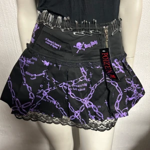 Kjol fr Poizen S - Kort svart kjol med lila kedjemönster, spetskant och säkerhetsnålar i midjan. Dragkedja framtill och detaljer med tryckta texter och dödskallemotiv. Märke Poizen. Storlek S, helt ny.